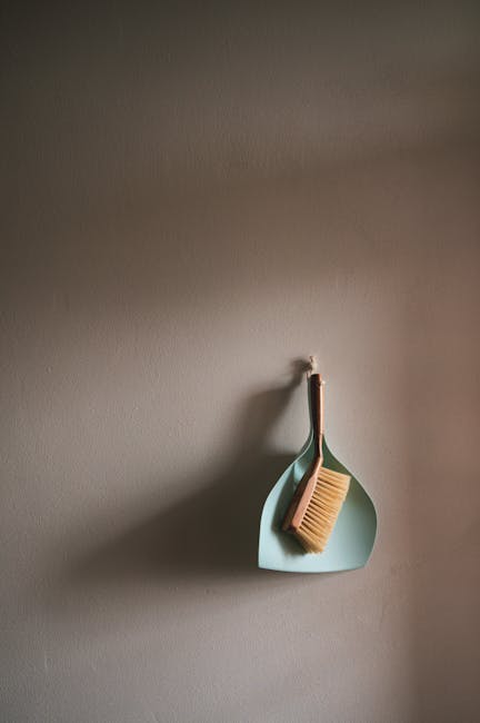 A light blue, modern wall hook attached to a neutral, beige wall holds a wooden-handled scrub brush with natural bristles and a matching wooden-handled cloth, arranged neatly. The wall surface appears