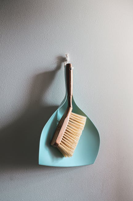 A person dressed in blue jeans and a grey top is using a blue mop with a white mop head to clean a wooden floor in a residential kitchen. The focus is on the mop and the wood surface, which appears cl
